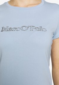 Světle modré tričko s krátkým rukávem, na kterém je mírně opotřebovaný černý logotyp "Marc O'Polo" přes hruď, vyrobené z měkké bavlny.