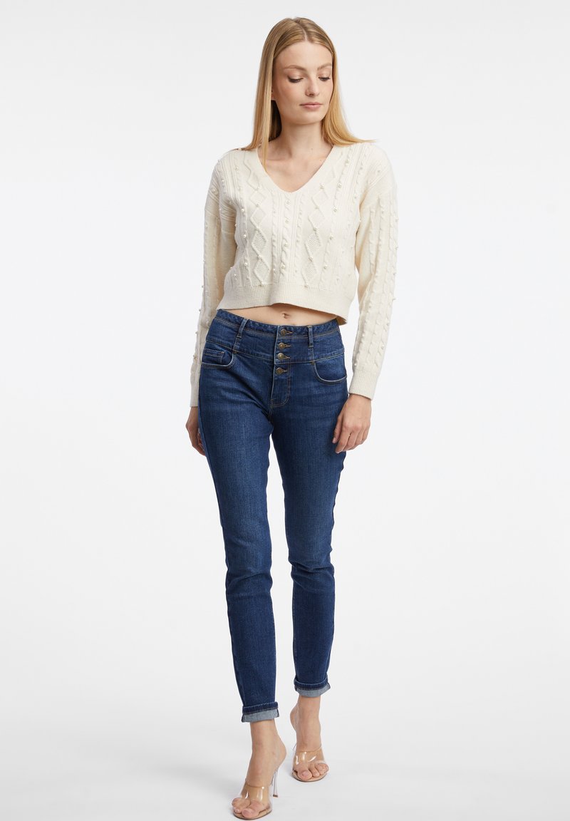 Pull cropped en tricot fantaisie de couleur crème avec un décolleté en V. Associé à un jean taille haute bleu foncé avec cinq boutons et des ourlets roulés. Talons clairs.
