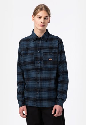 Camisa de franela para hombre en cuadros azul oscuro y negro. Cuenta con un cierre frontal de botones y dos bolsillos en el pecho con un pequeño parche de logo. Diseño de cuello clásico.