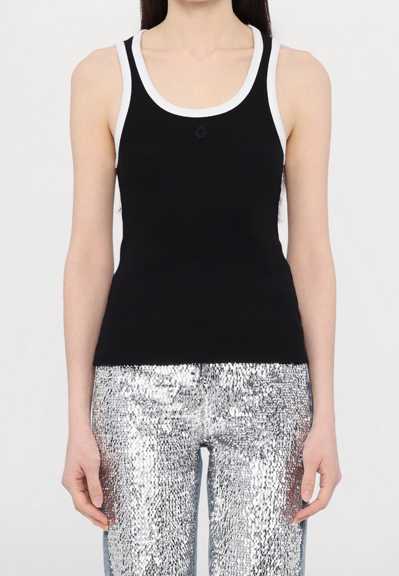 Femme portant un haut noir sans manches avec des bordures blanches et un pantalon argenté à sequins, debout devant un fond clair uni.