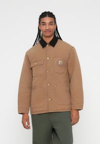 Brun utilityjakke med en sort kordfløjlskrave, fire frontlommer, metalknapper og et logo-patch på venstre bryst.