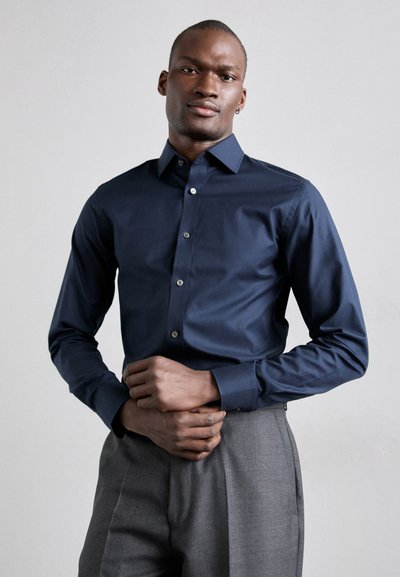 Chemise boutonnée bleu marine en tissu lisse avec des manches longues, col classique et boutons argentés, assortie à un pantalon gris.