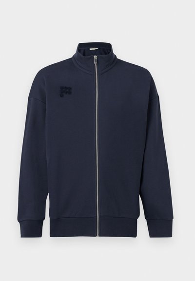 Veste zippée bleu marine avec col montant, manches longues, poignets et ourlet côtelés, et logo texturé discret sur le côté gauche de la poitrine.