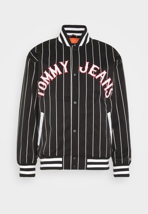 Jachetă bomber cu dungi negre și text roșu "TOMMY JEANS". Are mansete și guler rib, nasturi tip snap și buzunare laterale cu accente albe.
