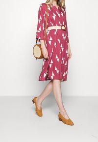 En rosa klänning med vita triangelmönster, trekvartslånga ärmar, midjebälte, ihop med vävda bruna skor och en rund brun väska.