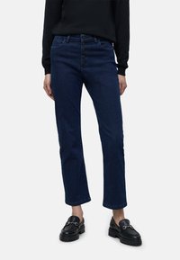 Jeans de mezclilla azul oscuro con pierna recta, cierre de cinco botones, costuras sutiles y un corte clásico, combinados con mocasines negros.