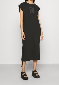 Czarna bawełniana sukienka do kolan z krótkimi rękawami i bocznymi rozcięciami. Posiada stonowane logo na klatce piersiowej i jest zestawiona z masywnymi czarnymi sandałami.