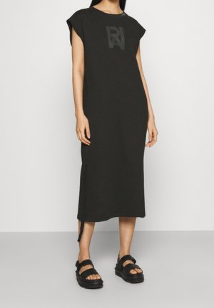 Robe noire en coton, longueur genoux, à manches courtes et fentes latérales. Présente un logo ton sur ton sur la poitrine et est assortie à des sandales noires épaisses.