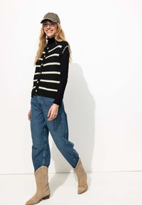 Gilet en maille rayé noir et blanc avec boutons, associé à un col roulé noir, un jean bleu et des bottes de cow-boy beiges. Une casquette texturée complète le look.