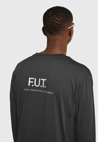 Osoba o krótkich włosach i kolczyku, ubrana w czarną bluzkę z długim rękawem z napisem "F.U.T. SICAL EDUCATION & TRAINING" na plecach.