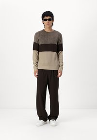 Maglione lavorato a maglia con strisce alternate beige e marrone scuro, texture a costine, abbinato a pantaloni larghi marroni e sneakers bianche.