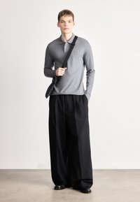 Graues Langarm-Polo mit Kragen und Logodetail, kombiniert mit lockeren schwarzen Hosen. Das Model trägt eine schwarze Umhängetasche und steht vor einem hellen Hintergrund.