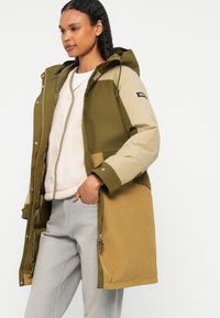 Parka w kolorze blokowym w oliwkowej zieleni i beżu, z kapturem, zapięciem na zamek błyskawiczny oraz miękką podszewką. Zawiera kieszenie i luźny krój.