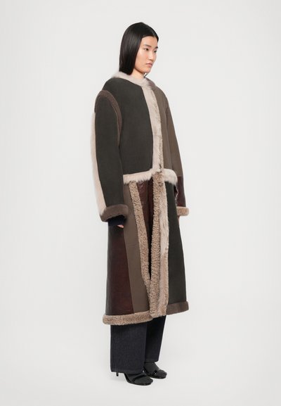 Palton lung din blană de oaie maro, cu o margine de blană moale, având texturi variate, conceput cu un design deschis în față și o croială supradimensionată, asortat cu pantaloni negri.