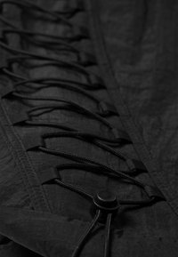 Black textile featuring laces along one edge, with a smooth texture and a toggle fastener. Minimalistic design with no visible patterns.