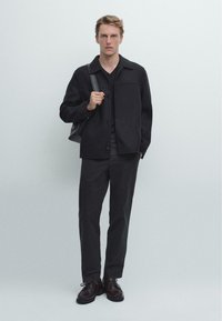 Černý krátký kabát z vlněné směsi se zapínáním na knoflíky, v kombinaci s černými kalhotami a hnědými koženými botami. Černá kabelka přes rameno dodává celý vzhled.