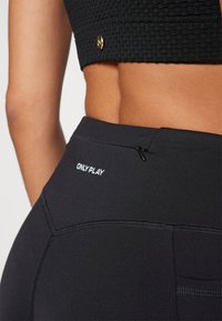 Leggings noirs avec une taille haute, dotés d'une poche zippée sur le côté et d'un logo "ONLYPLAY" blanc au dos. Texture lisse et design ajusté.