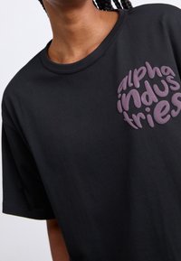 Camiseta de algodón negra con un logotipo elevado en forma de círculo de color púrpura que dice "alpha industries" en el lado izquierdo. Diseño de mangas cortas y cuello redondo.