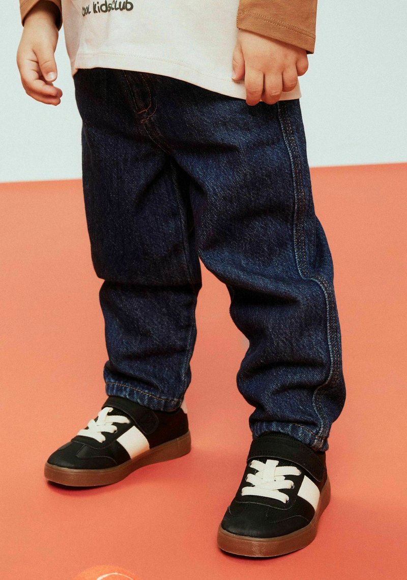 Dark blue denim jeans with a relaxed fit and prominent stitching, paired with black sneakers featuring a white stripe and laces.