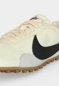 Λευκό αθλητικό παπούτσι Nike με μαύρο σήμα, μπεζ κορδόνια, υφασμένη σόλα από καουτσούκ και ραμμένες λεπτομέρειες σε απλό φωτεινό φόντο.