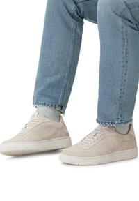 Baskets en suède beige clair à profil bas, avec un détail de laçage et une semelle texturée, accompagnées d'un jean en denim bleu clair et de chaussettes gris côtes.