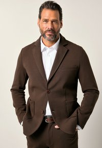 Blazer marrone con una vestibilità su misura, rever a punta, due tasche anteriori e chiusura con un solo bottone, indossato sopra una camicia bianca.