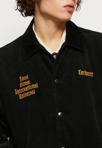 Czarna kurtka z sztruksu z żółtym haftowanym napisem na klatce piersiowej. Charakterystyczne detale to duży kołnierz i zatrzaski.