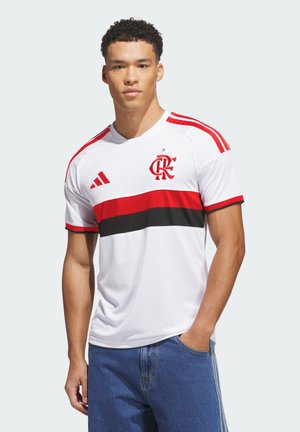 Męski model noszący biały sportowy jersey z czerwonymi i czarnymi poziomymi paskami, czerwonym logo i paskami na ramionach, w połączeniu z niebieskimi dżinsami.