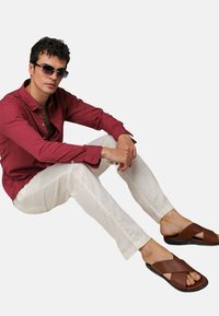 Camicia rossa con texture, pantaloni in lino bianchi e sandali marroni con incrocio. Modello seduto, con occhiali da sole, che mostra un abbigliamento estivo casual.