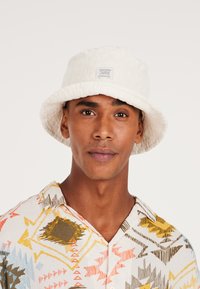 Witte bucket hat van een gestructureerde stof met een gestikte label; gedragen met een bedrukte, kortesleeve, kraagshirt in levendige kleuren.