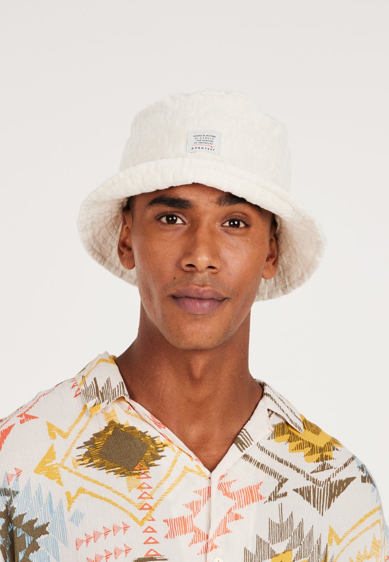 Witte bucket hat van een gestructureerde stof met een gestikte label; gedragen met een bedrukte, kortesleeve, kraagshirt in levendige kleuren.