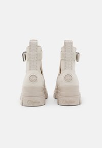 Bottes à plateforme beige épaisses avec une texture lisse, dotées d'une sangle de marque et d'un détail de logo, découpes sur les côtés, et boucle réglable.