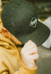 Casquette noire avec une texture mouchetée, arborant un patch logo blanc en forme de diamant. La visière est plate et sombre, avec des détails de couture.