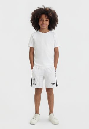 Göndör hajú gyermek áll előre nézve, egyszerű fehér pólót, fehér logós rövidnadrágot és fehér tornacipőt visel, világos háttér előtt.