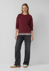 Vrouw staat in een casual outfit met een bordeauxrode trui over een gestreept shirt, wijde zwarte jeans en beige sneakers tegen een effen achtergrond.
