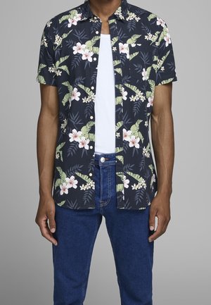 Mand iført mørkeblå kortærmet skjorte med hvidt og grønt blomstermønster over hvid T-shirt og blå jeans foran grå baggrund.