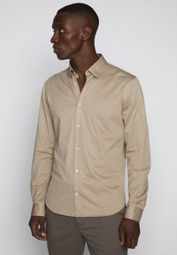 Beige langärmliges Hemd aus glattem Stoff mit spitzem Kragen, Vorderknopfverschluss und Manschettenärmeln, das ein tailliertes Design zeigt.