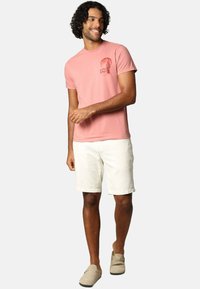 T-shirt rose clair avec un motif graphique sur la poitrine gauche, associée à un short blanc et des chaussures slip-on beiges. Texture lisse et décontractée.