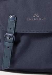 Tamsiai mėlynas audeklo kuprinis krepšys su odine juostele ir spaudžiamu mygtuku, ant kurio iškilmingai užrašyta "DOUGHNUT" logotipas virš kalnų piešinio.