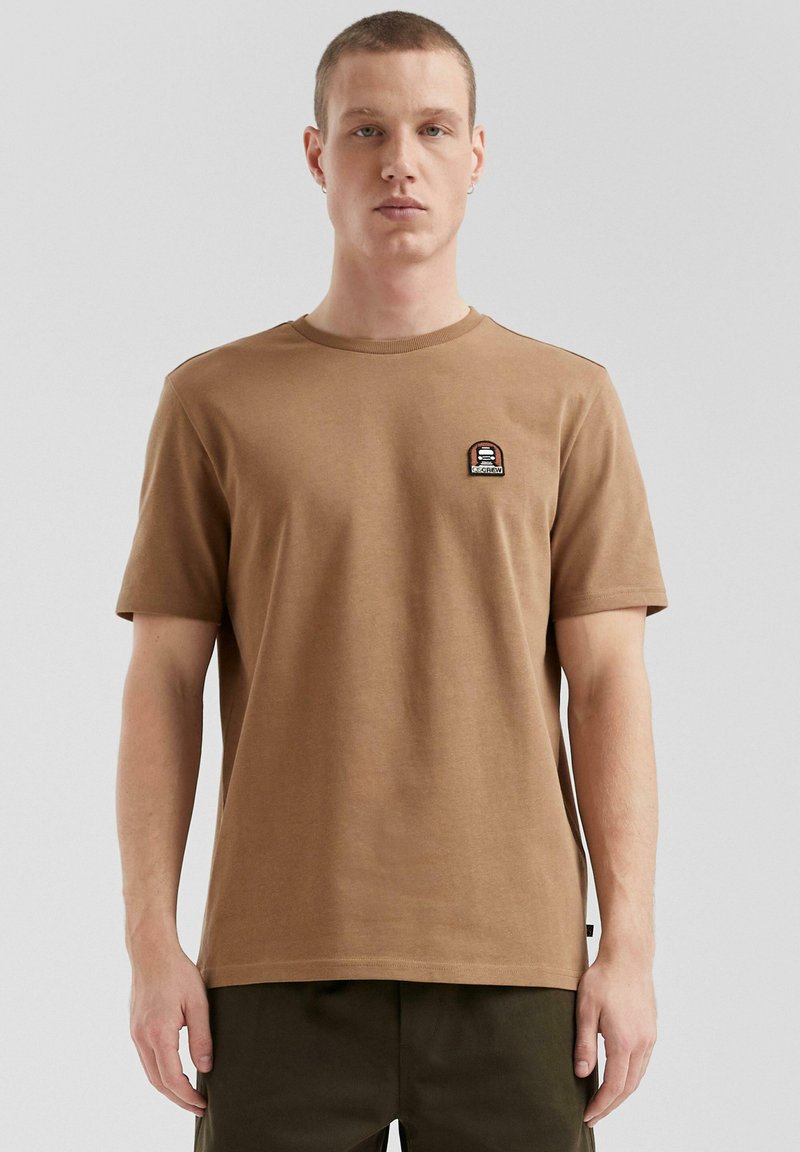 T-shirt in cotone marrone con colletto tondo, maniche corte e un piccolo ricamo sul lato sinistro del petto. Tessuto liscio, vestibilità comoda.