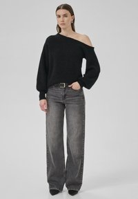 Overdimensioneret sort sweater med off-shoulder design, tekstureret stof, parret med højtaljede grå brede jeans og sorte spidse sko.