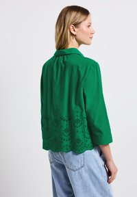 Blouse verte avec un col, des manches trois-quarts et des découpes élaborées sur la moitié inférieure. Fabriquée en tissu léger.