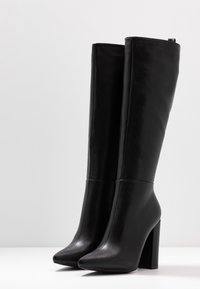 Para wysokich czarnych skórzanych butów na obcasie w kształcie bloku oraz ze spiczastymi noskami, stojących pionowo na białej powierzchni na jednolitym tle.