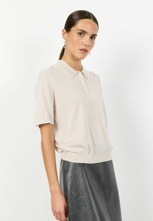 Sötét hajú nő alacsony copfban, gyöngyfülbevalóval, rövid ujjú, bézs csíkos galléros pólóban és fekete bőr szoknyában, oldalra fordulva áll.