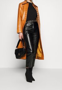 Donna che indossa un cappotto arancione con motivo coccodrillo, pantaloni neri a vita alta con motivo coccodrillo, top nero, tiene in mano una borsa nera e indossa stivali neri con tacco.