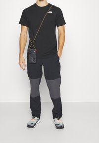 Black t-shirt with logo, gray and black cargo pants with knee patches, gray sneakers with orange accents, carrying a small black pouch.