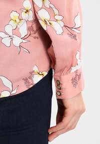 Blus med långt ärm i mjuk rosa tyg med stora vita och gula blommönster; knappdetaljer på ärmsluten; slät textur.