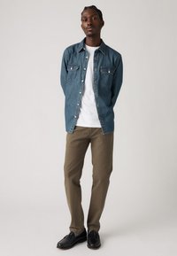 Denim button-up shirt in blue with two chest pockets, paired with a white t-shirt and olive green pants. Black loafers complete the outfit.
