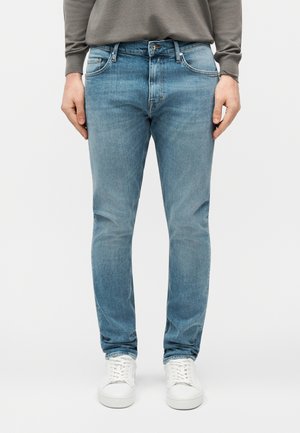 Hombre vistiendo vaqueros ajustados azul claro y zapatillas blancas, de pie contra un fondo liso.