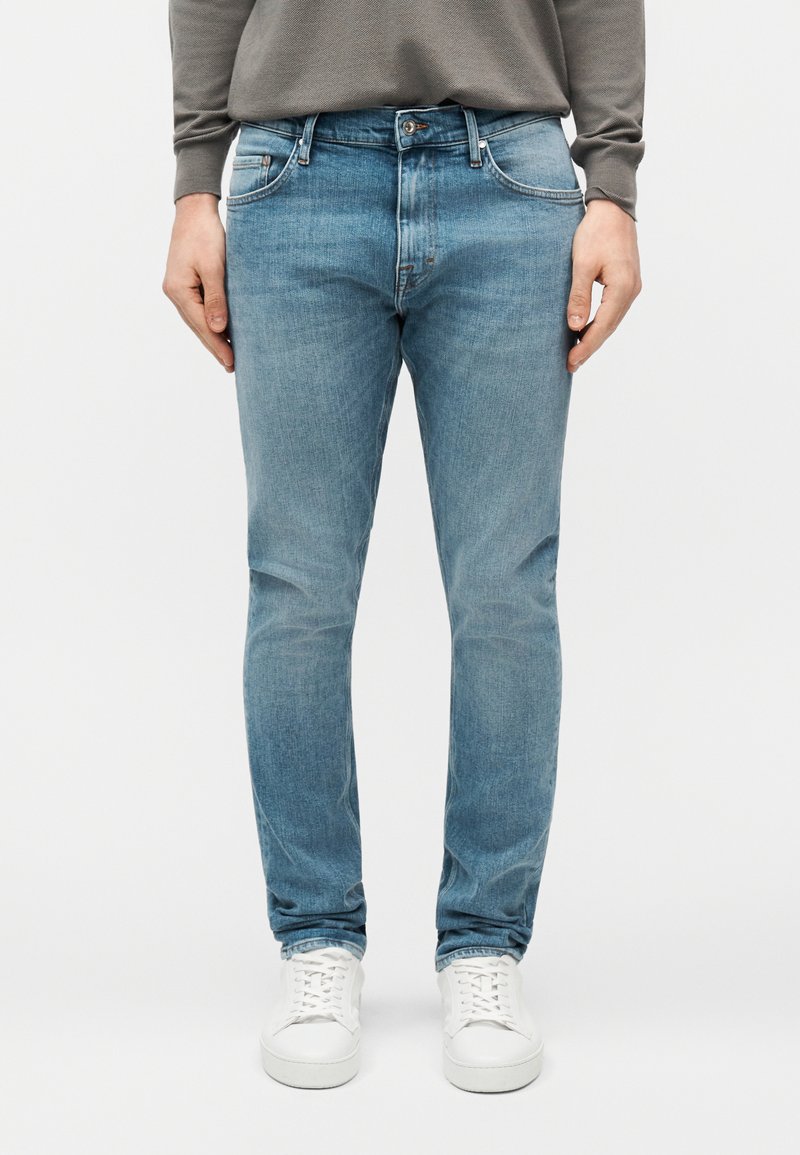 Άνδρας που φοράει ανοιχτό μπλε τζιν στενής γραμμής και λευκά αθλητικά παπούτσια, στέκεται μπροστά σε απλό φόντο.